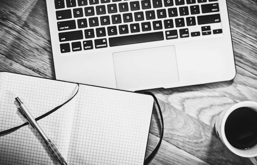 Black and white image of a workspace featuring a laptop, notebook, pen, and coffee cup.